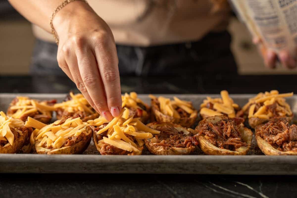 Hollowed potato skins loaded with pulled pork, being sprinkled with shredded cheese.