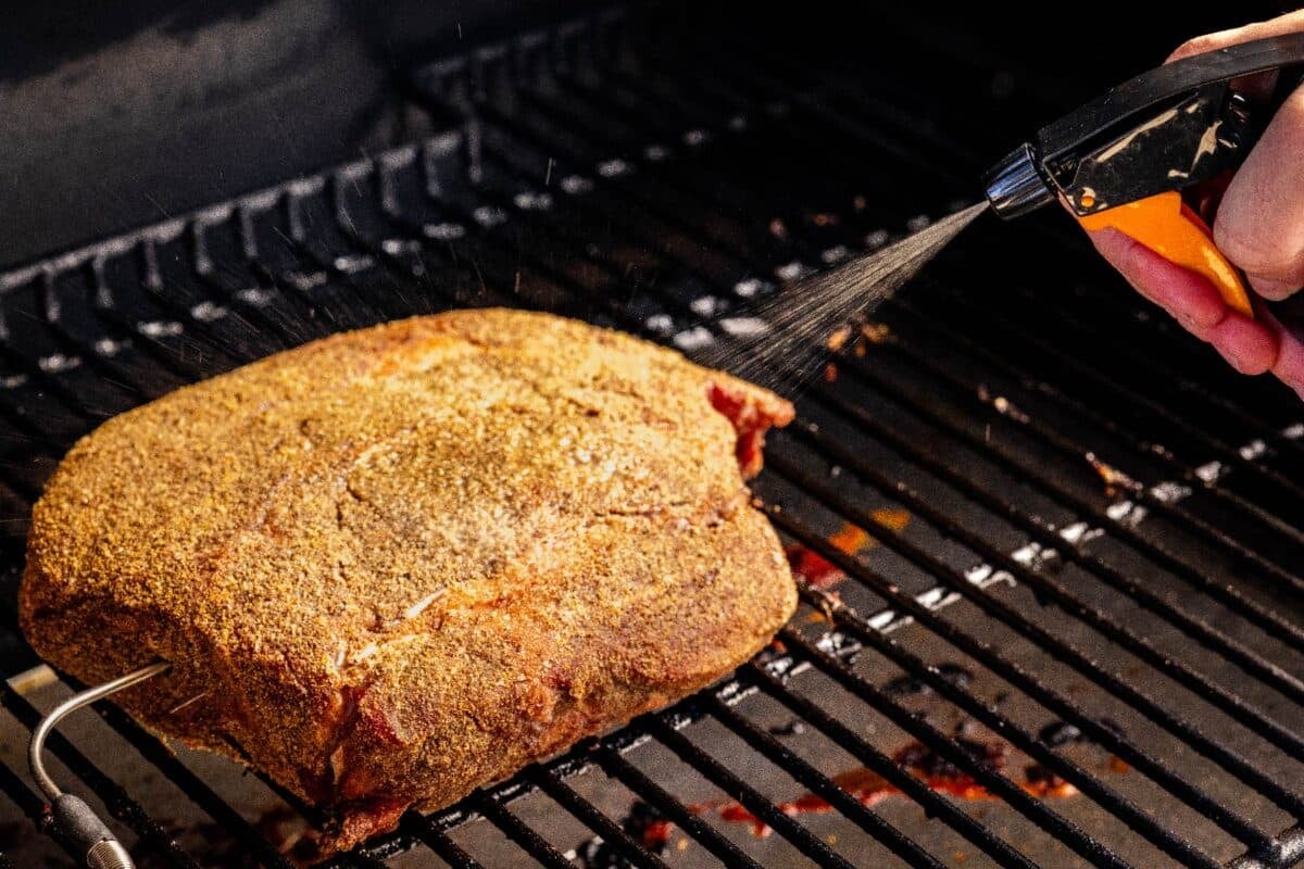 Seasoned chuck roast sitting on smoker grates being spritzed with stock.