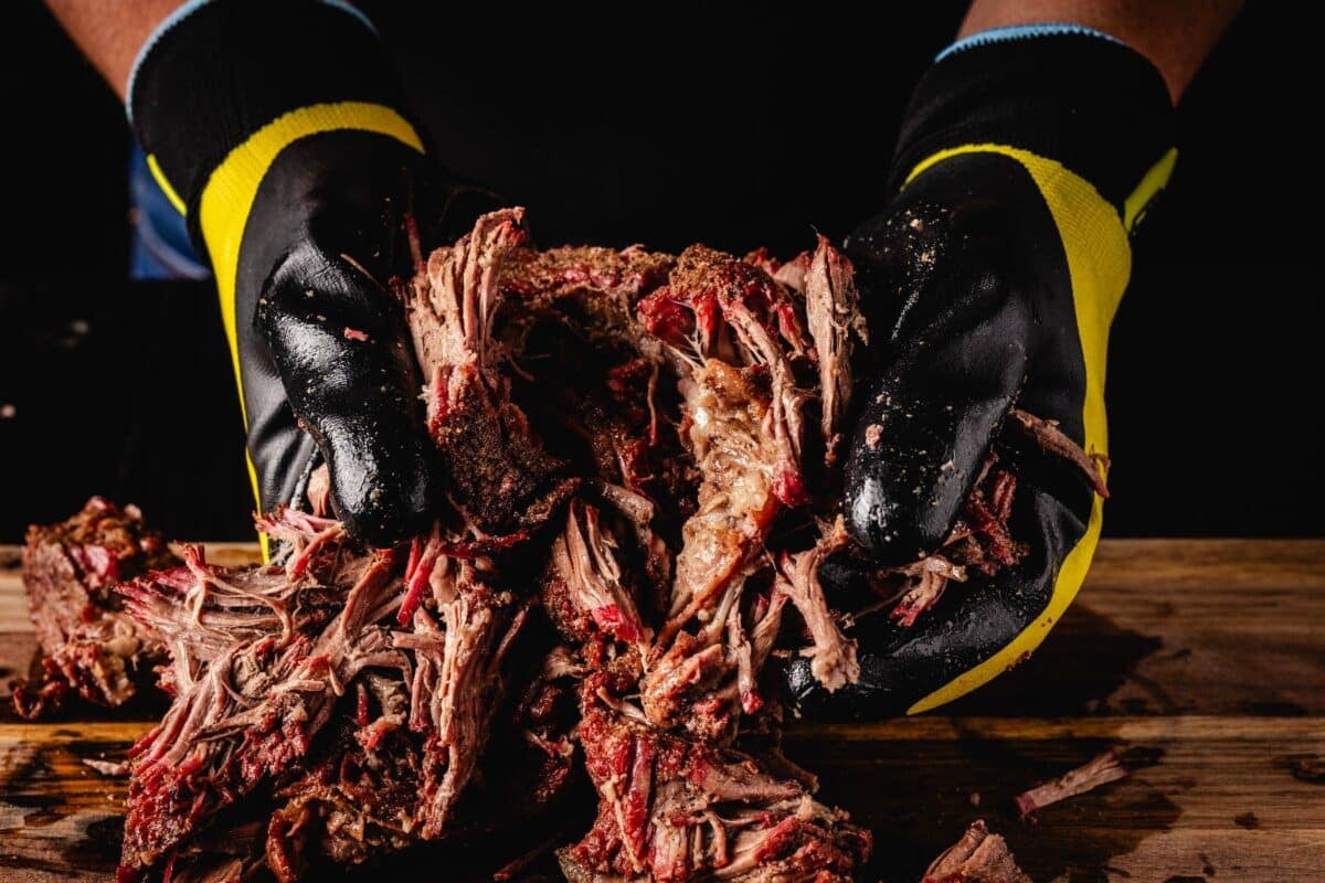 Gloved hands pulling beef on a wooden cutting board.