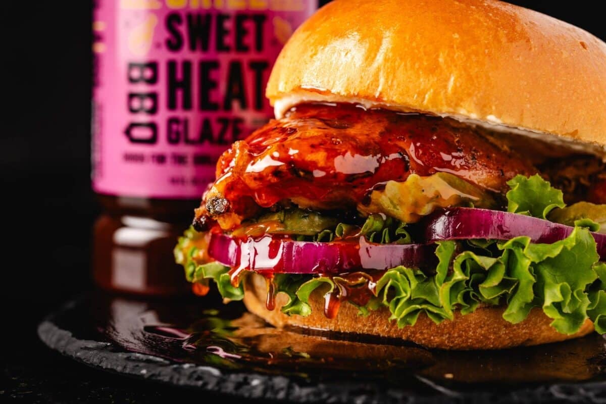 A hot chicken sandwich with lettuce, sliced onion, and BBQ sauce, in front of a bottle of sauce.