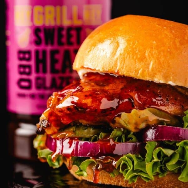 A chicken sandwich with lettuce, sliced onion, and BBQ sauce, in front of a bottle of sauce.