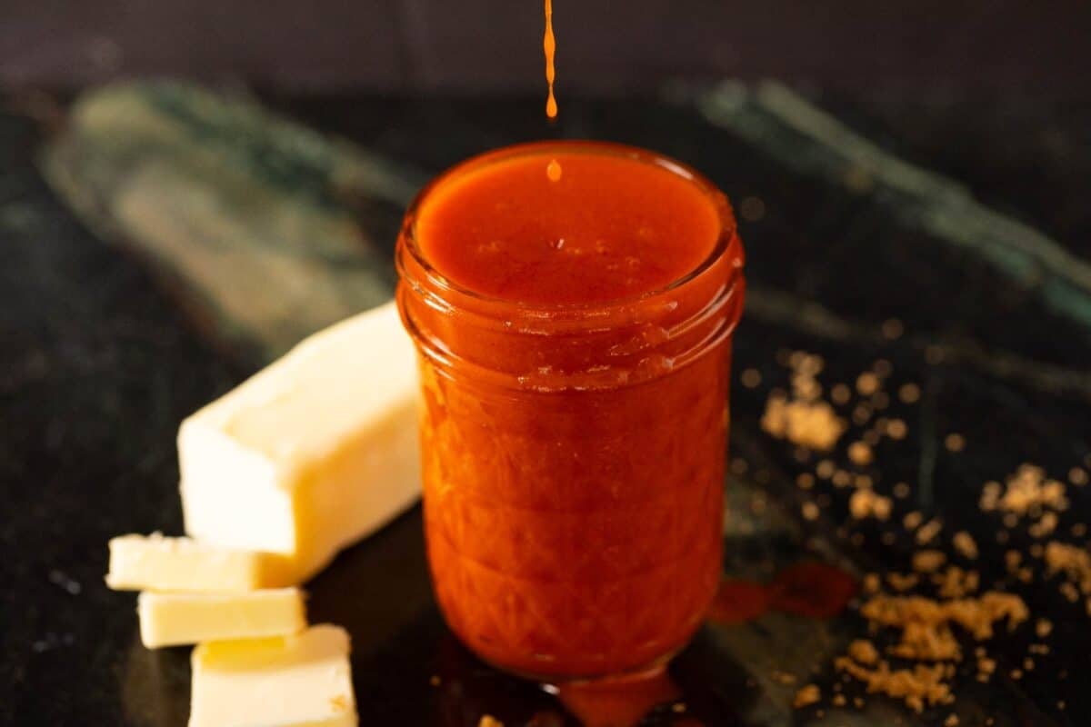 A clear mason jar with buffalo sauce, next to a stick of partially sliced butter.