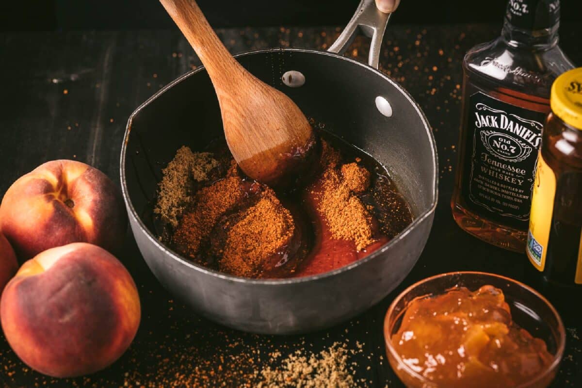 Sauce ingredients in pot being stirred with wooden spoon, next to peaches and other ingredients.