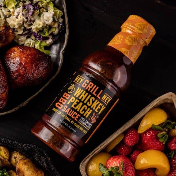 A bottle of Whiskey Peach BBQ Sauce next to a bowl of chicken and coleslaw, and a dish or fruit.