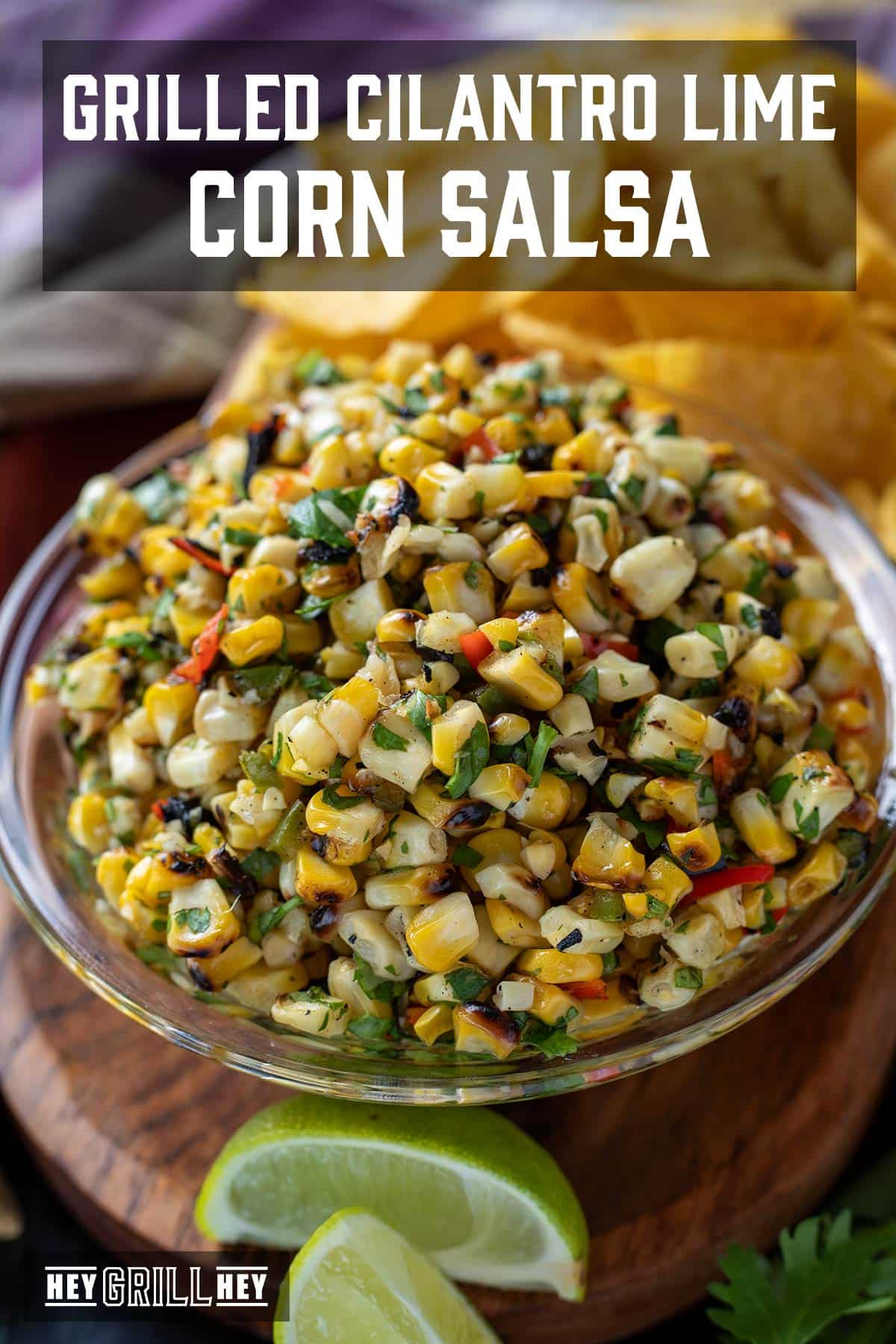 Corn salsa in a glass bowl next to a lime wedge and pile of tortilla chips. Text overlay reads "Grilled Cilantro Lime Corn Salsa."