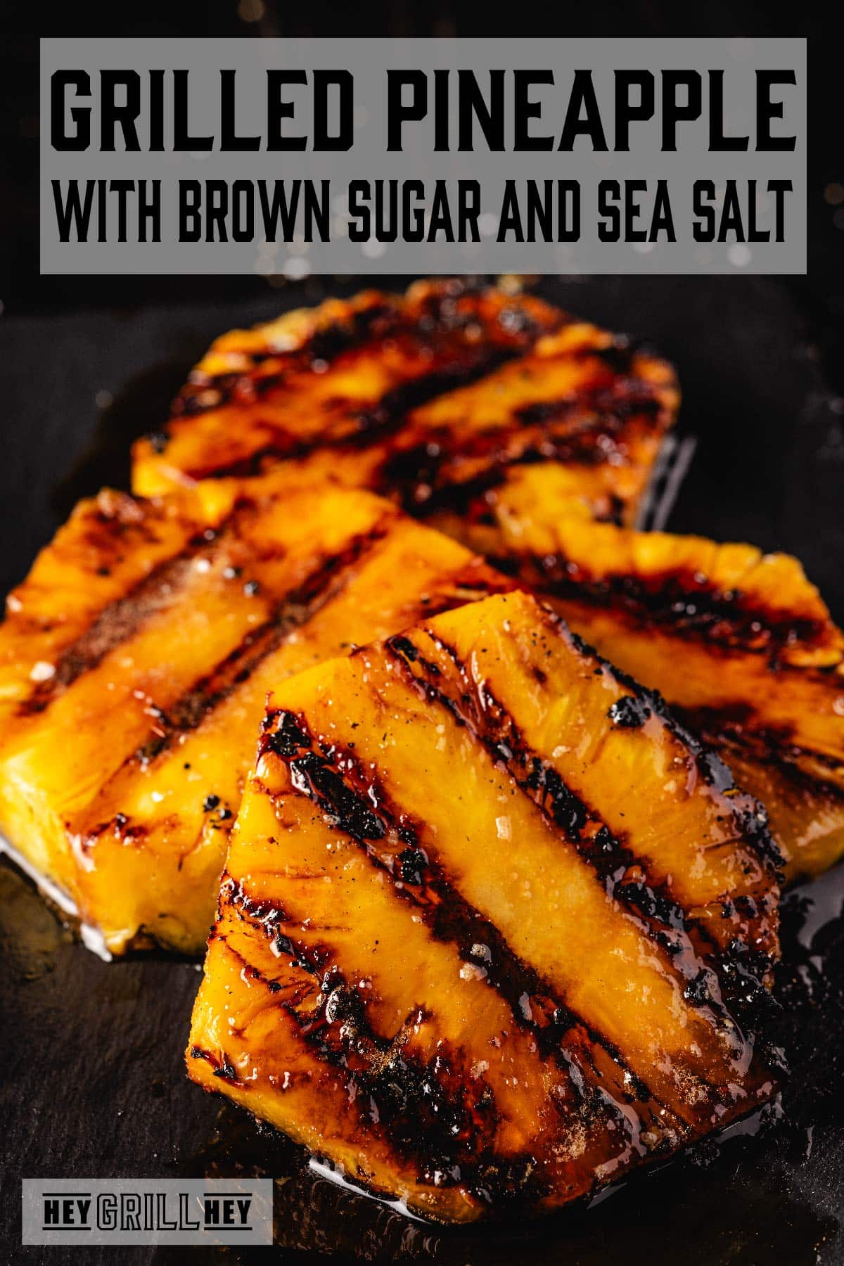 Grilled pineapple slices stacked on a black countertop. Text reads "Grilled Pineapple with Brown Sugar and Sea Salt."