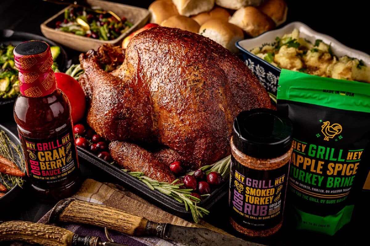 A smoked turkey on a table next to rolls and other sides, and packages of BBQ seasonings and sauces.