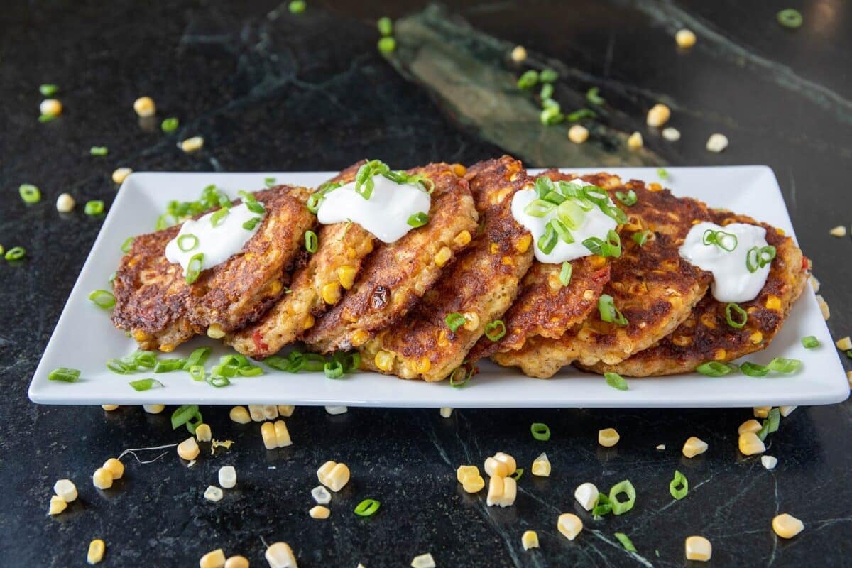 Fried corn fritters on white serving platter on countertio, sprinkled with corn and green onions.