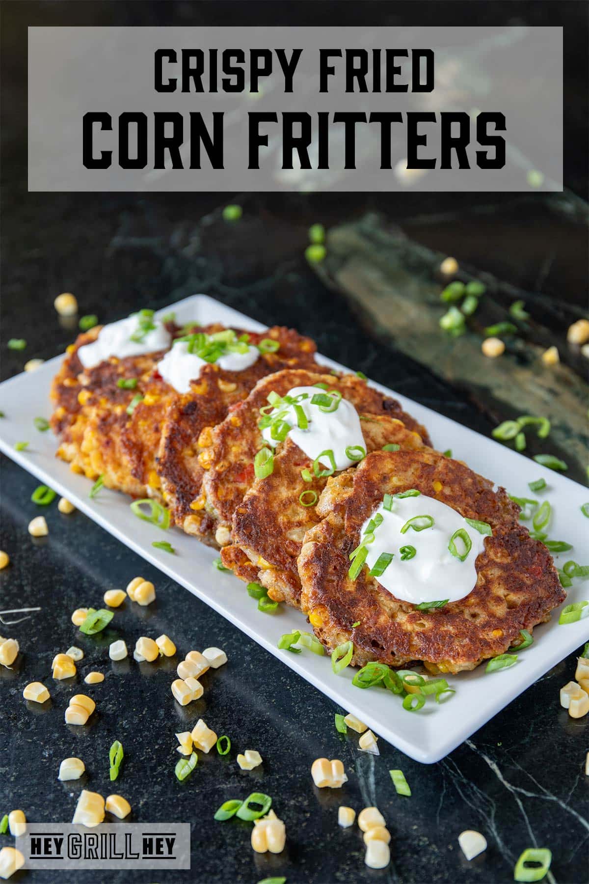 Fried corn fritters on a white serving dish, garnished with sliced green onions, and surrounded by loose corn kernels. The text overlay reads "Crispy Skillet Corn Fritters."