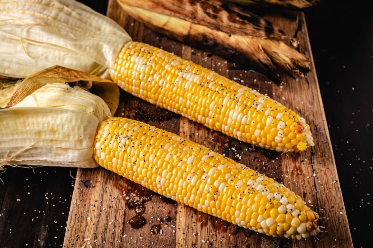 Corn with husks still attached on a wood cutting board.