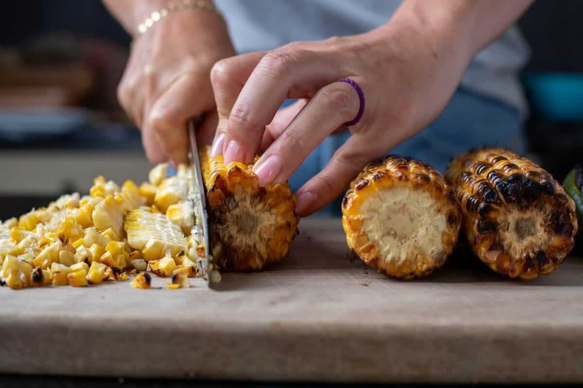 Grilled earns of corn on a cutting board. A hand holding a knife is slicing the kernels off one.