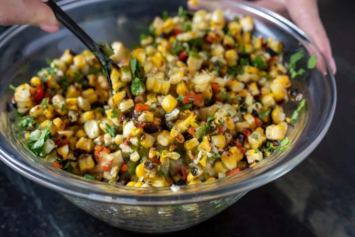 A spoon mixing corn salsa in a glass bowl.