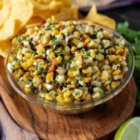 Corn salsa in a glass bowl on a wooden platter with tortilla chips and a lime wedge.