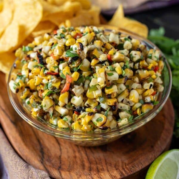 Corn salsa in a glass bowl on a wooden platter with tortilla chips and a lime wedge.