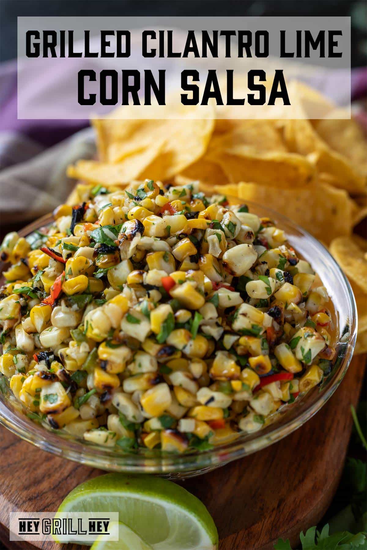 Corn salsa in a glass bowl next to a lime wedge and pile of tortilla chips. Text overlay reads "Grilled Cilantro Lime Corn Salsa."