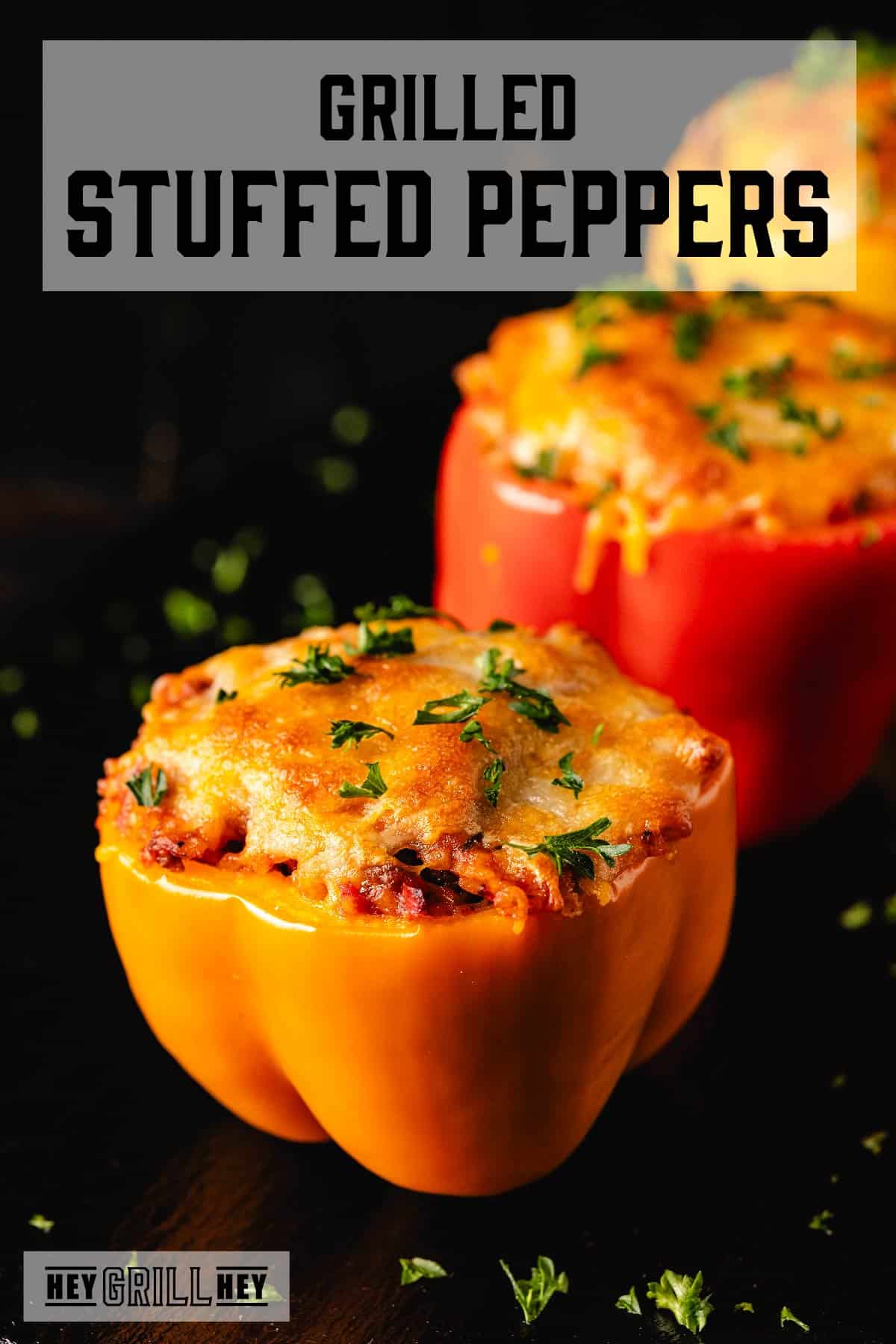 One red and one orange bell pepper half, both with filling on a black platter. The text overlay and the top reads "Grilled Stuffed Peppers," and the small one at the bottom reads "Hey Grill Hey."