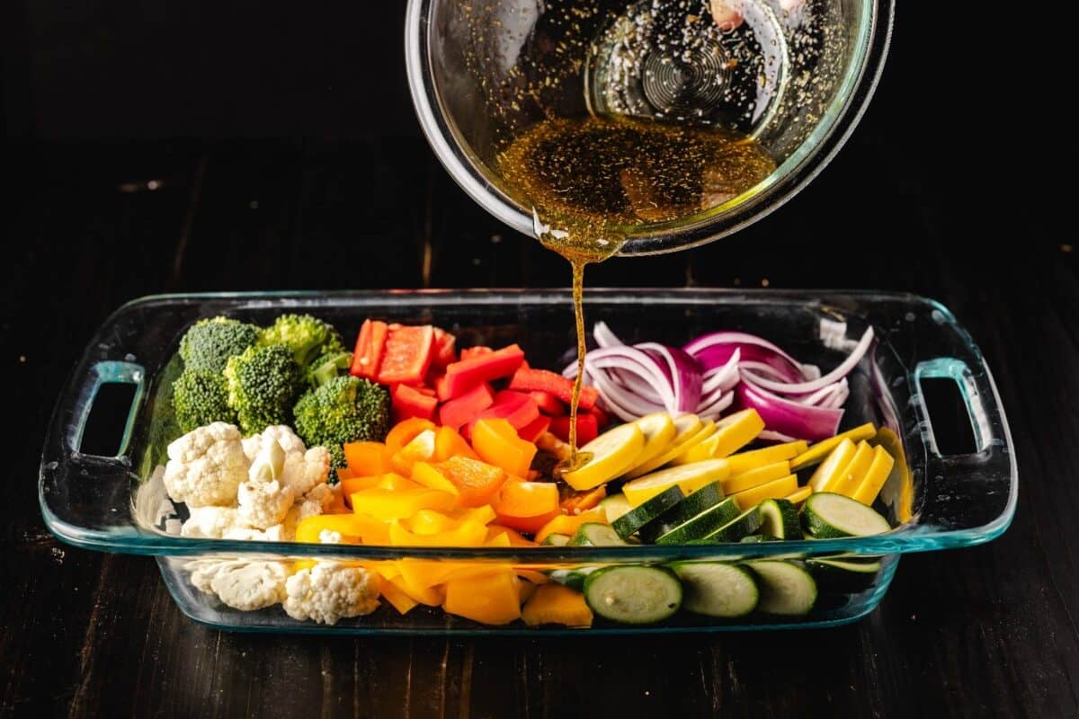 Broccoli, cauliflower, onions, and peppers in a baking dish being drizzled with olive oil.