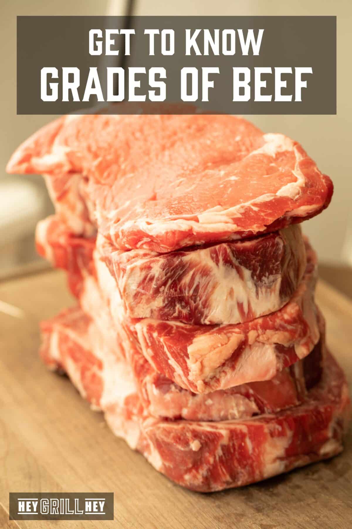 Raw steaks piled on a wooden cutting board. The text overlay reads "Get to Know Grades of Beef" at the top, and "Hey Grill Hey" at the bottom.