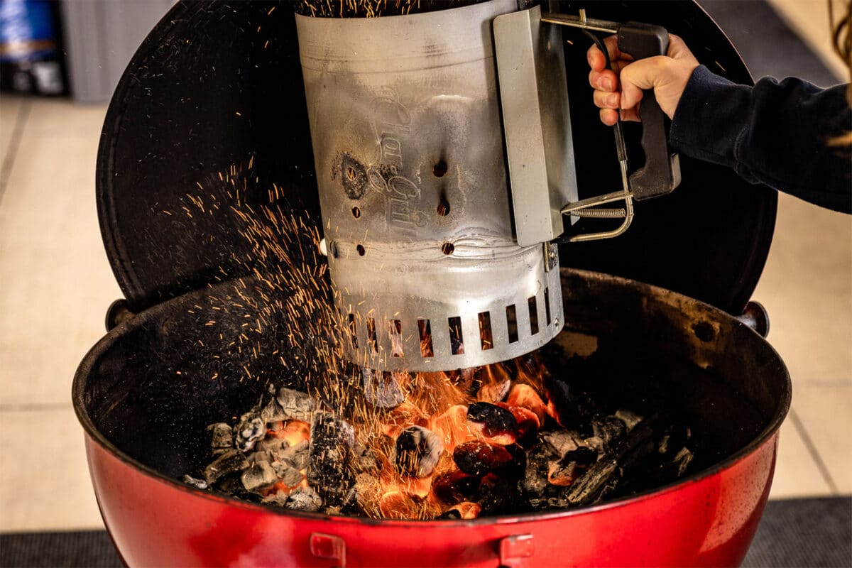 A silver charcoal chimney being emptied with flaming briquettes settling into the base of a charcoal grill.