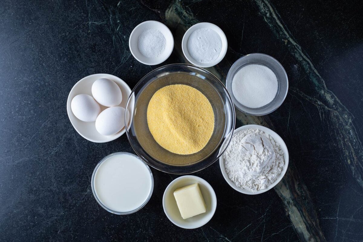 Cornmeal, flour, and other ingredients for corn muffins in glass bowls arranged on a marble countertop.