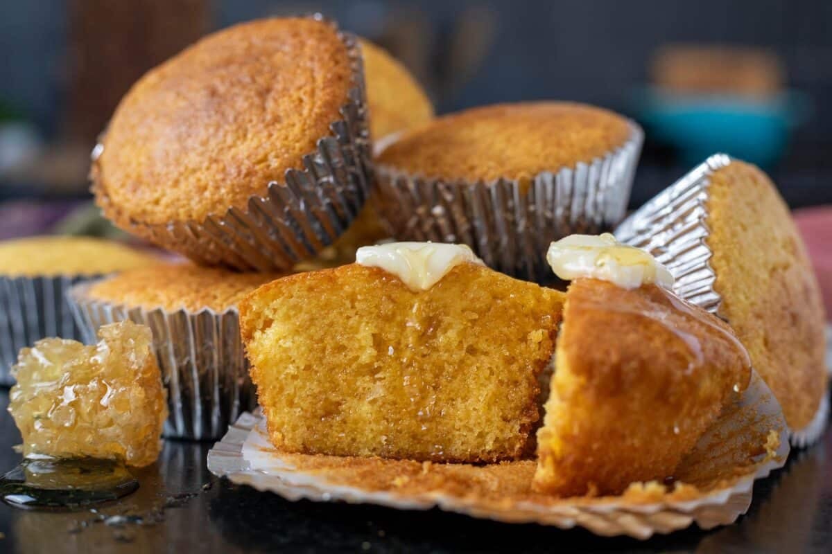 Corn muffins in paper liners piled behind one muffin that's sliced in half and topped with butter and honey.