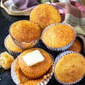 Corn muffins in liners, drizzled with honey, and stacked on a counter. One muffin is topped with a pad of butter.