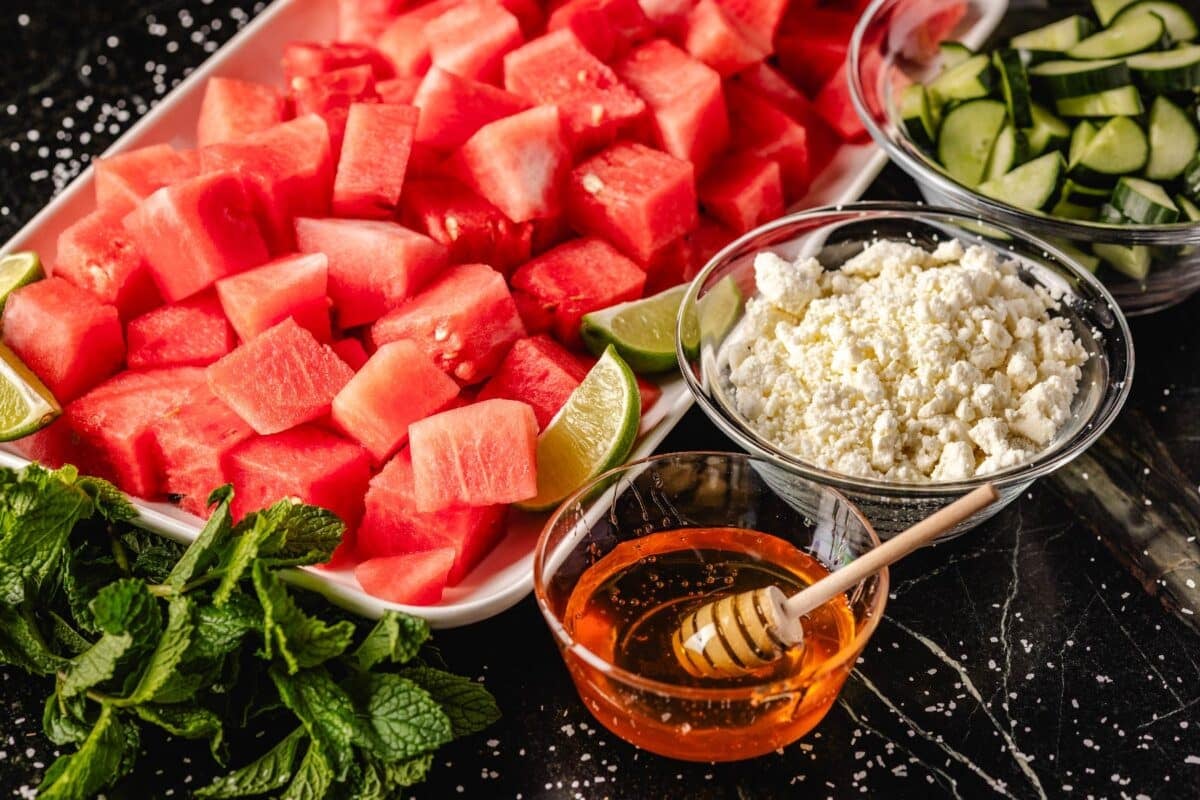 Sliced watermelon on a tray next to bowls of honey and feta crumbles, and fresh mint leaves.