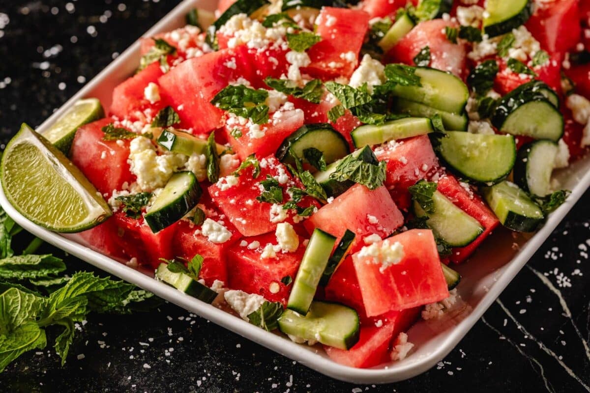 Sliced fruit topped with feta cheese and mint, next to lime wedges.