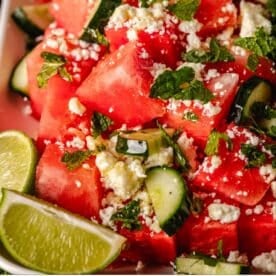 Sliced watermelon and cucumber topped with feta cheese and mint, next to lime wedges.