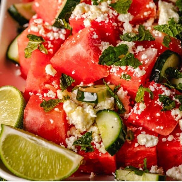 Sliced watermelon and cucumber topped with feta cheese and mint, next to lime wedges.