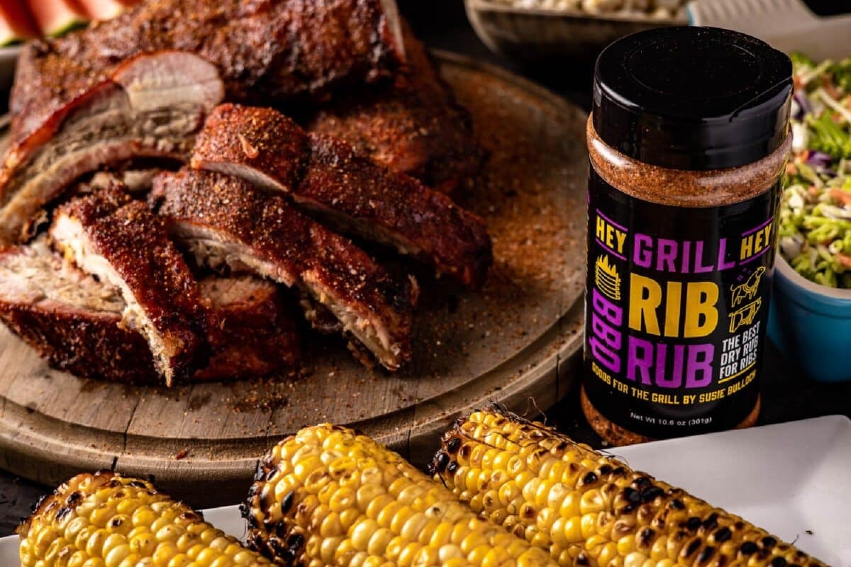 A bottle of BBQ seasoning next to a plate of ribs, a plate of corn, and other side dishes.