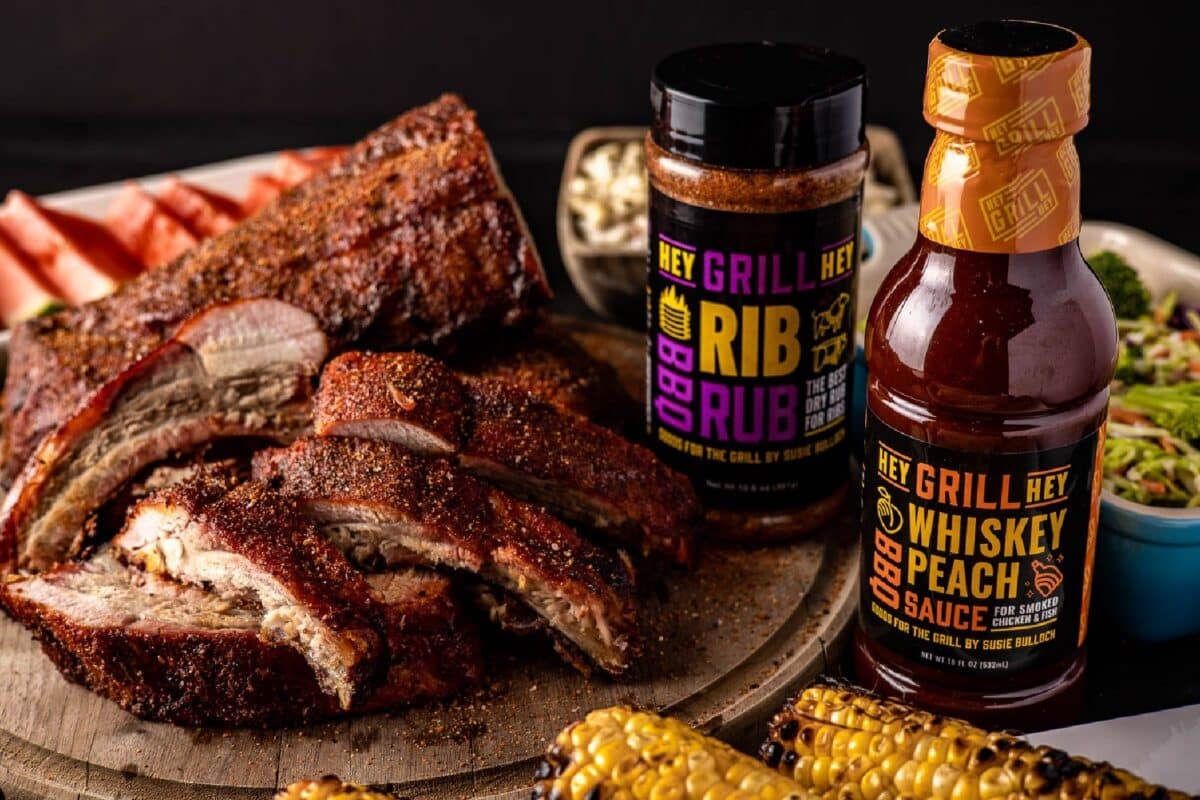 Bottles of BBQ condiments next to a plate of sliced ribs and two ears of grilled corn.