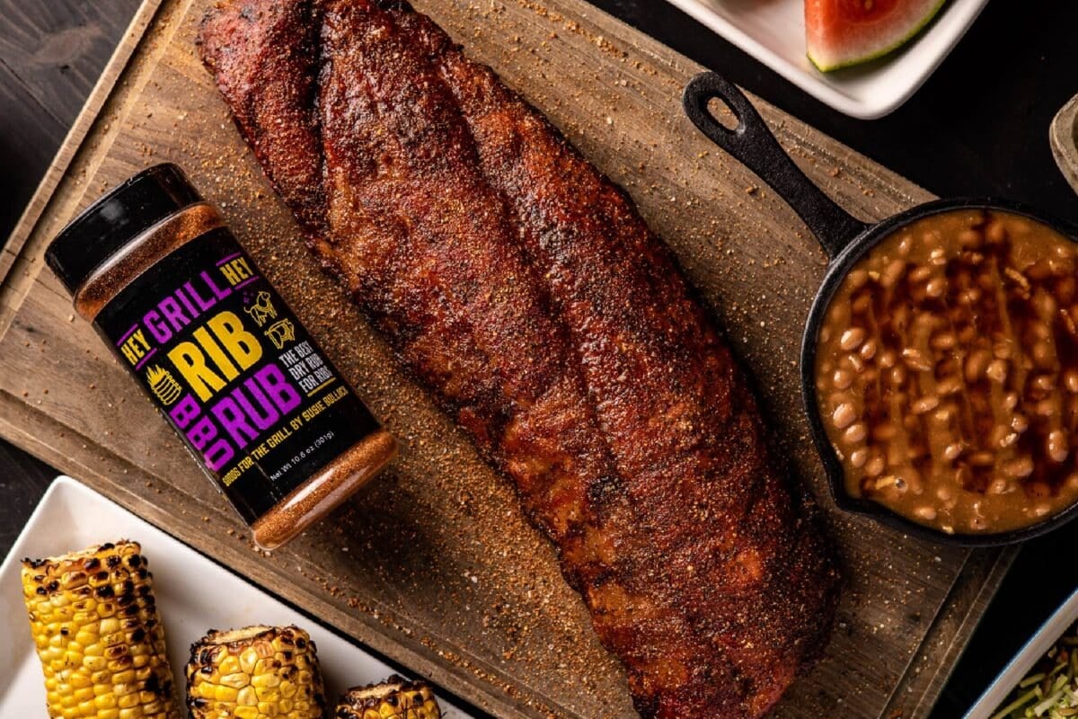 A rack of cooked, unsliced ribs, a skillet of beans, and a bottle of Rib Rub on a wooden cutting board.