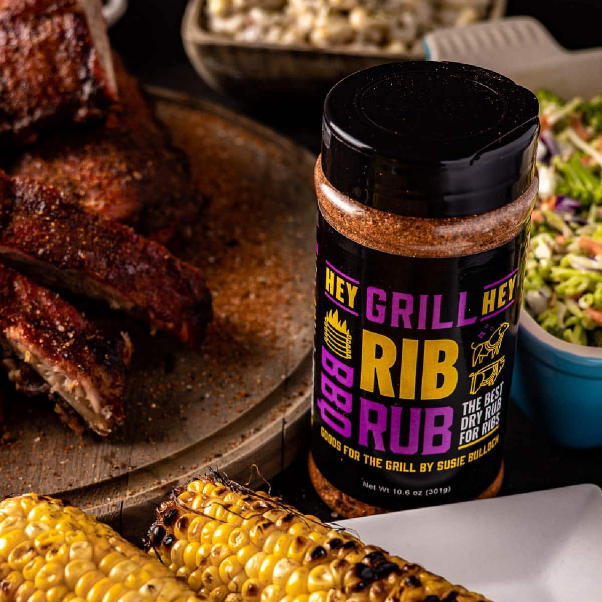 A bottle of BBQ seasoning next to a plate of ribs, a plate of corn, and other side dishes.