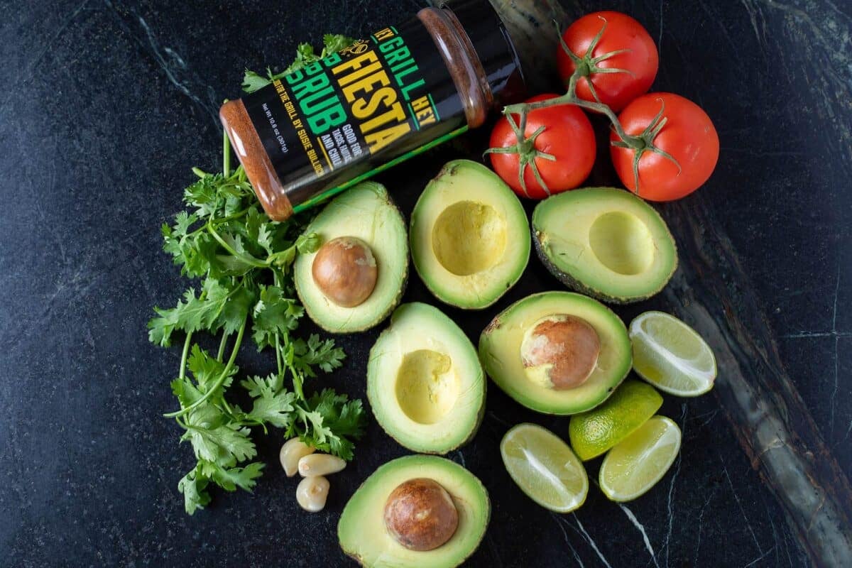 Avocado halves, tomatoes, cilantro, and a jar of seasoning on a tabletop.