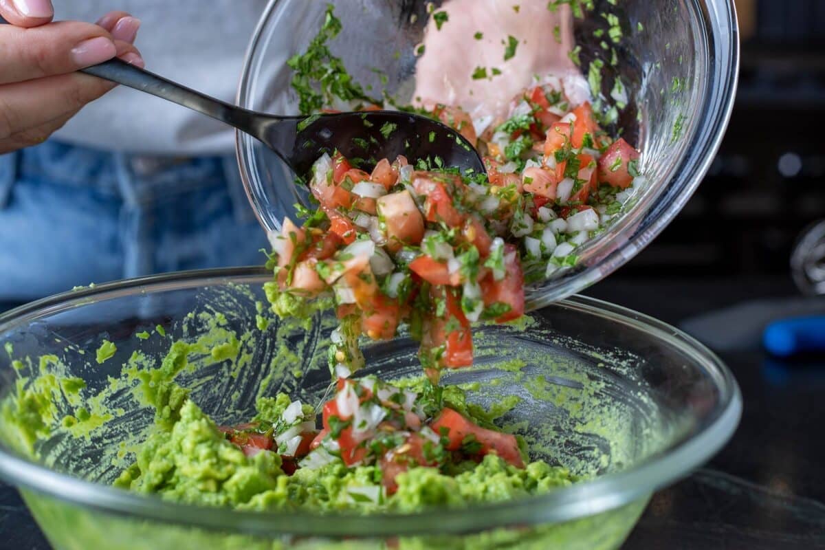 Diced tomatoes being scooped from a bowl with a spoon into a bowl of mashed avocado.