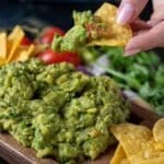 Fresh guacamole in a wooden serving dish with tortilla chips. A hand is holding a chip with a scoop of guacamole.