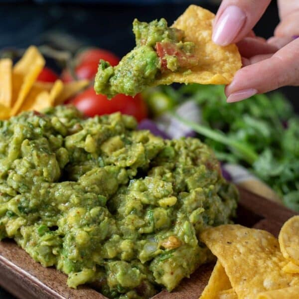 Fresh guacamole in a wooden serving dish with tortilla chips. A hand is holding a chip with a scoop of guacamole.