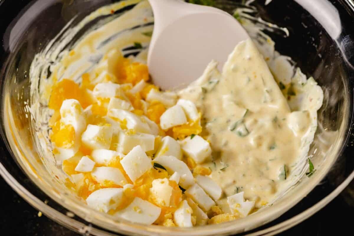 A rubber spatula mixing chopped eggs into a salad mixture in a glass bowl.