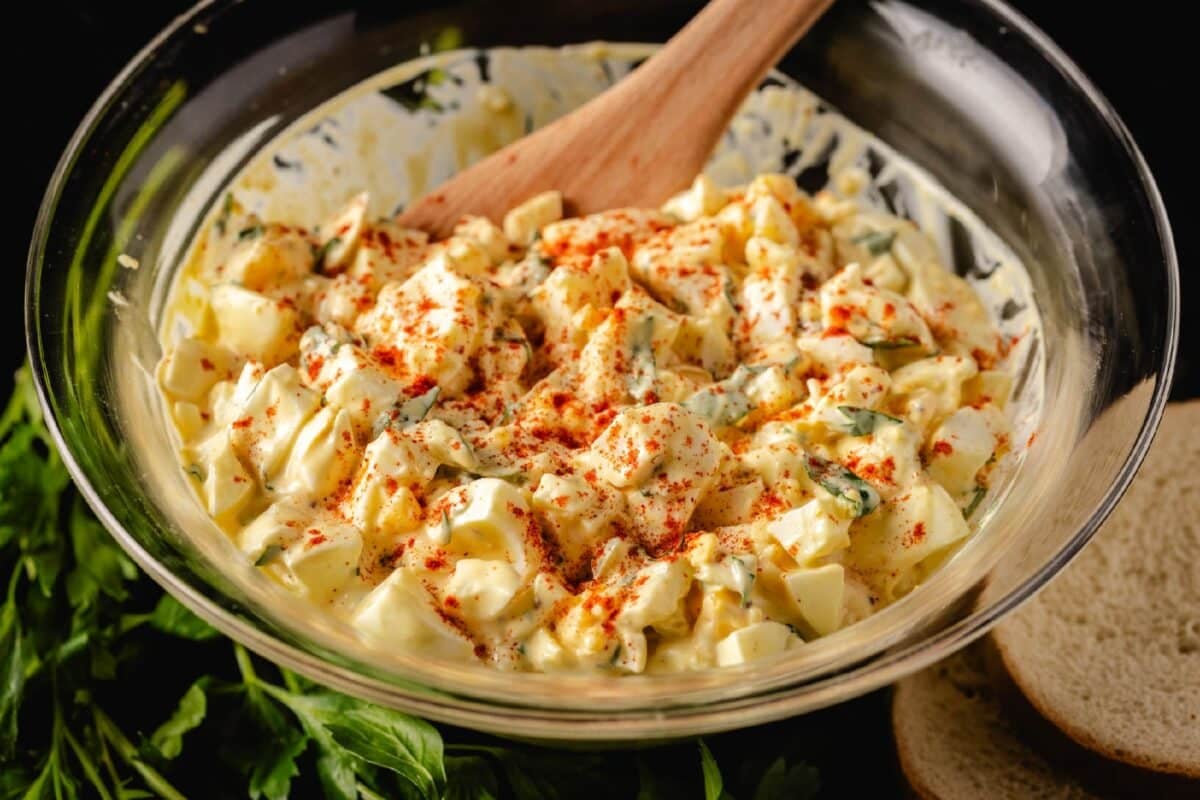 A wooden spoon stirring egg salad in a glass bowl.