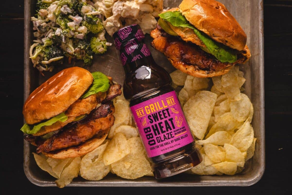 Bottle of Sweet Heat Glaze on a baking sheet with potato chips, chicken sandwiches, and broccoli salad.