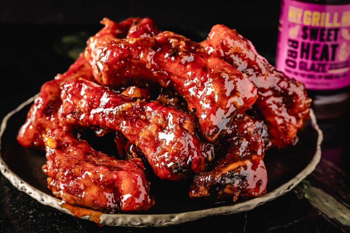 Glazed ribs piled on a black plate next to a bottle of Sweet Heat Glaze.