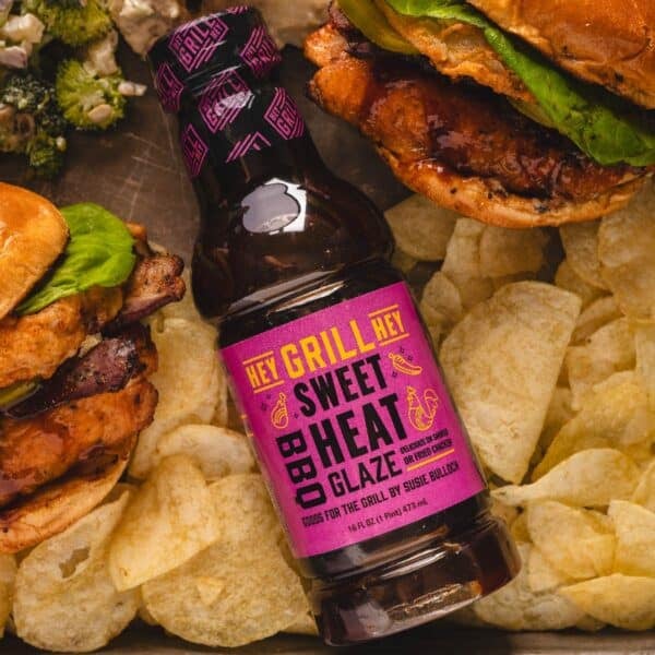 Bottle of Sweet Heat Glaze on a baking sheet with potato chips, chicken sandwiches, and broccoli salad.