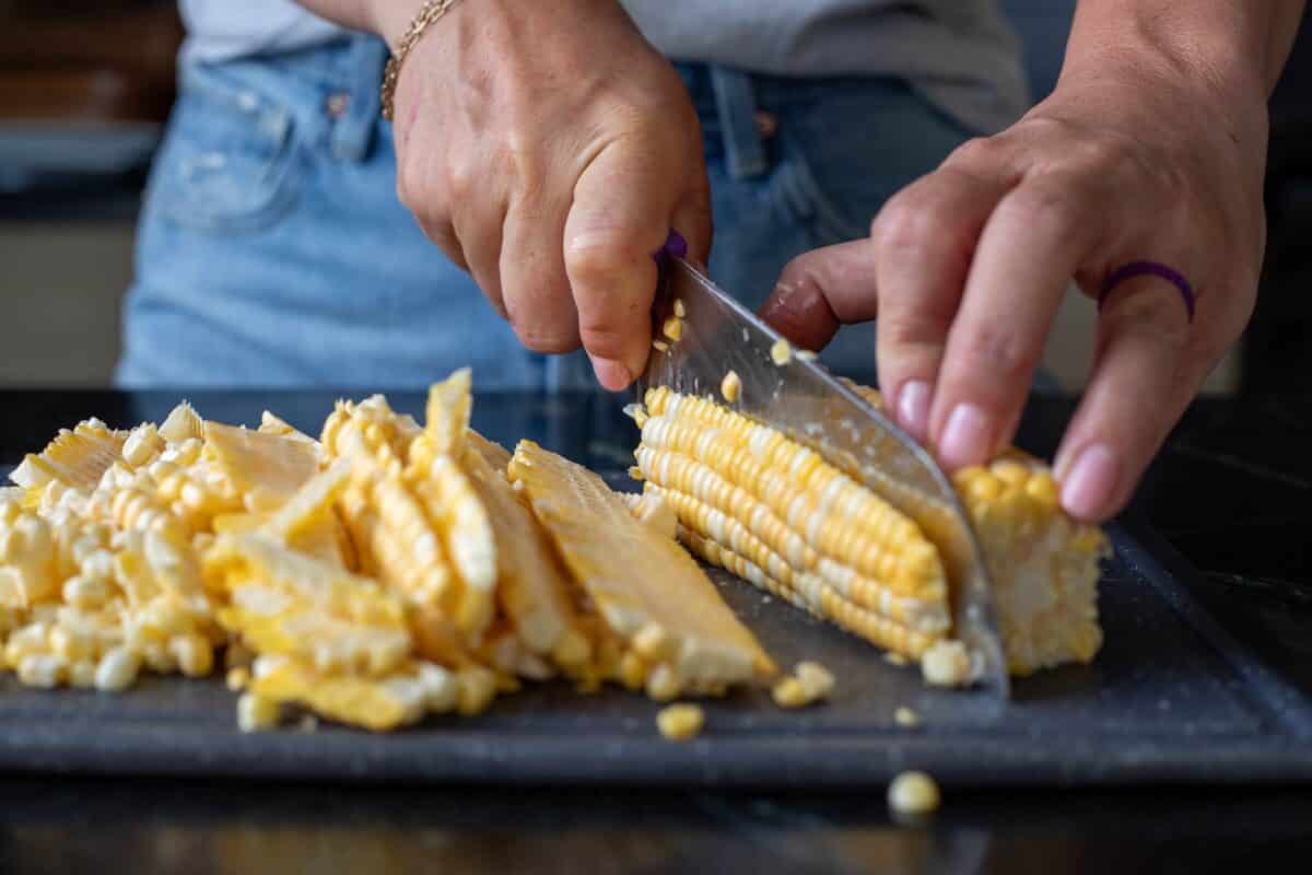 A knife chopping kernels of corn off a cob on a cutting board next to a pile of chopped kernels.