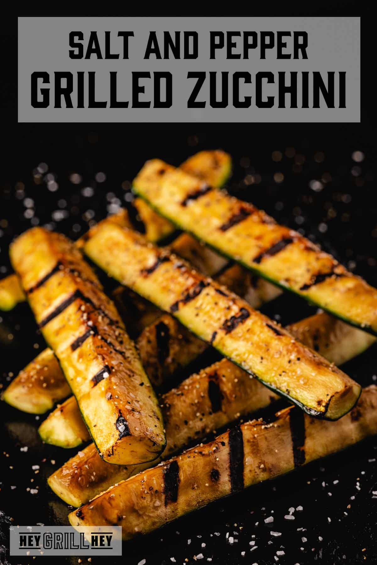 Zucchini spears with grill marks sprinkled with sea salt on a black serving platter. Text reads "Salt and Pepper Grilled Zucchini."