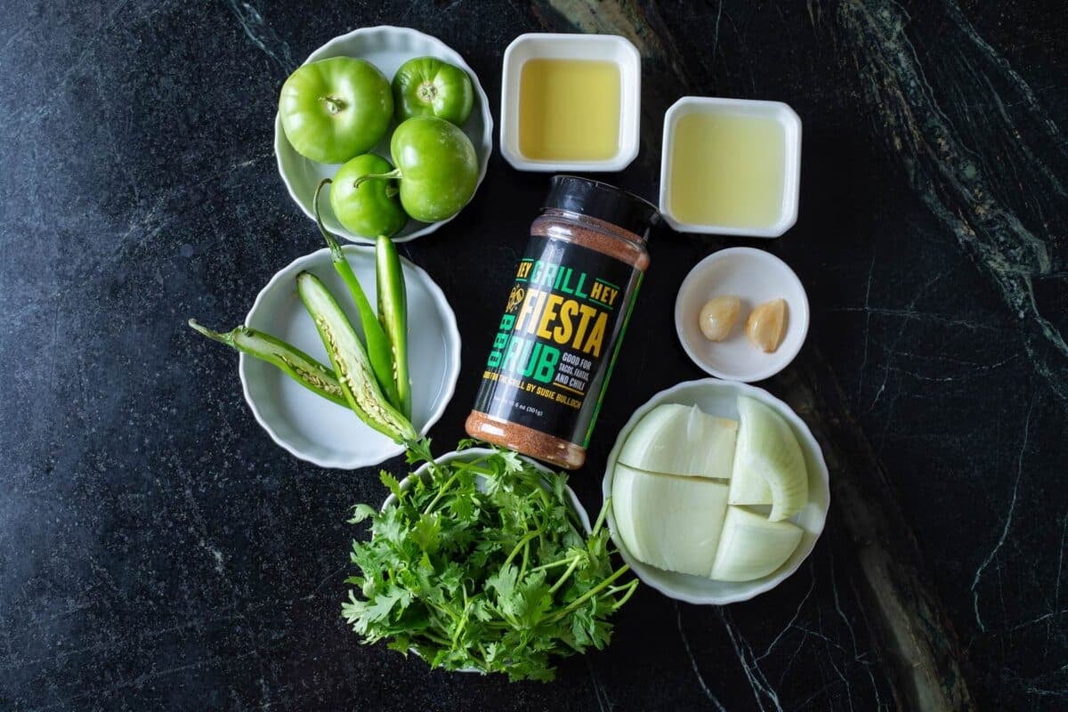 A bottle of Fiesta Rub on a table surrounded by white bowls with chiles, and other fresh ingredients.