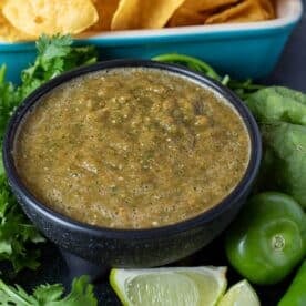 Smoked salsa in a black bowl garnished with cilantro leaves and lime wedges, next to a blue platter with chips.