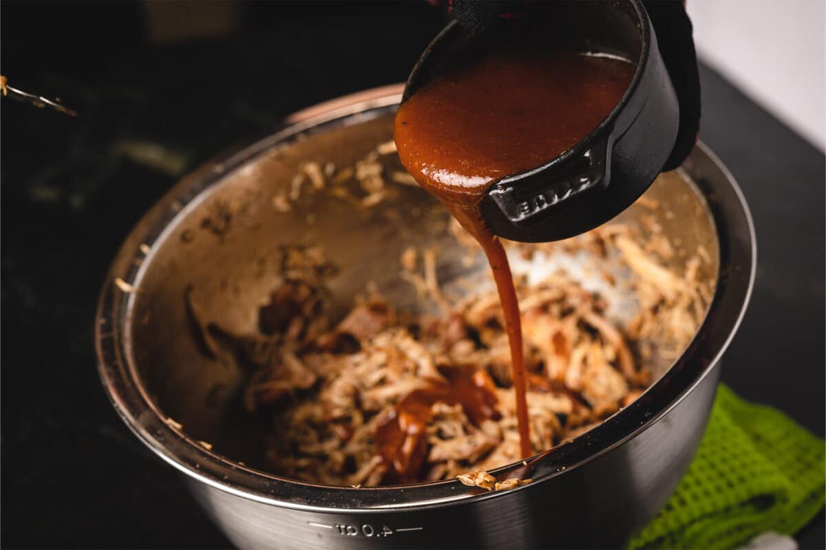 Red sauce being drizzled over shredded chicken in a silver bowl.