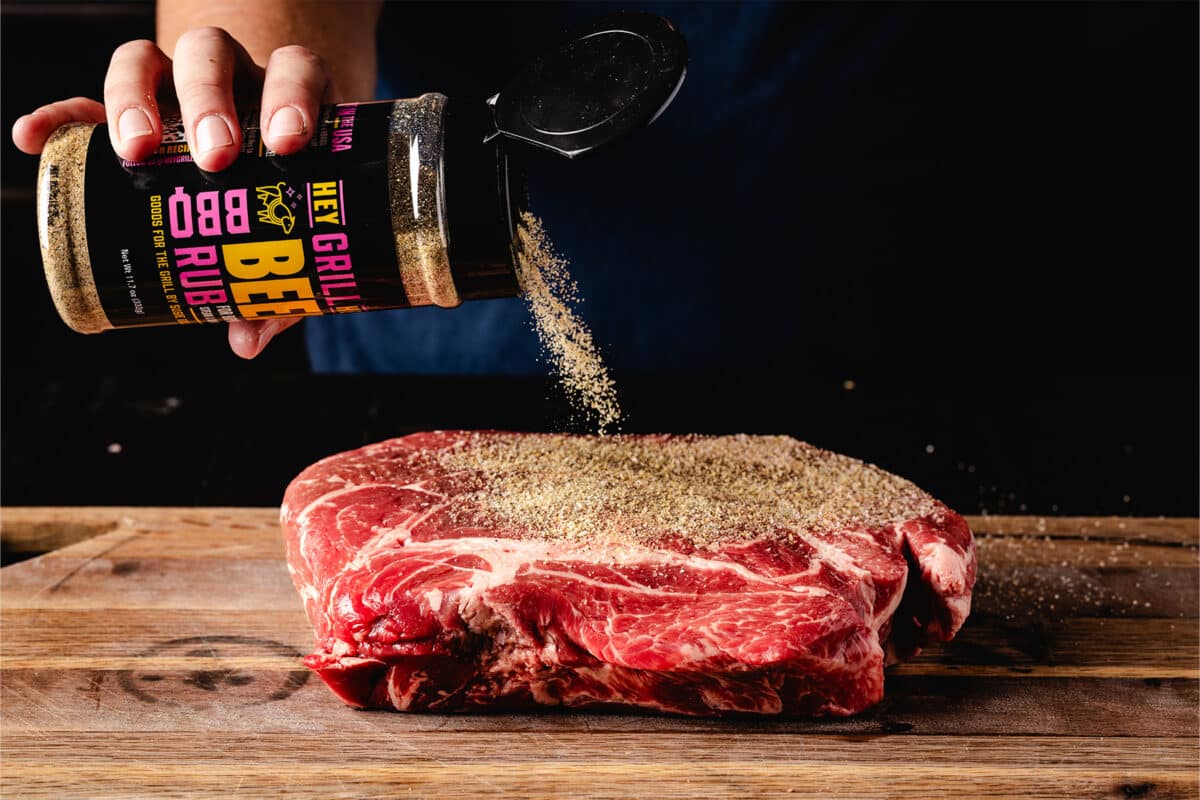 A large cut of steak being seasoned liberally with rub on a wooden cutting board.