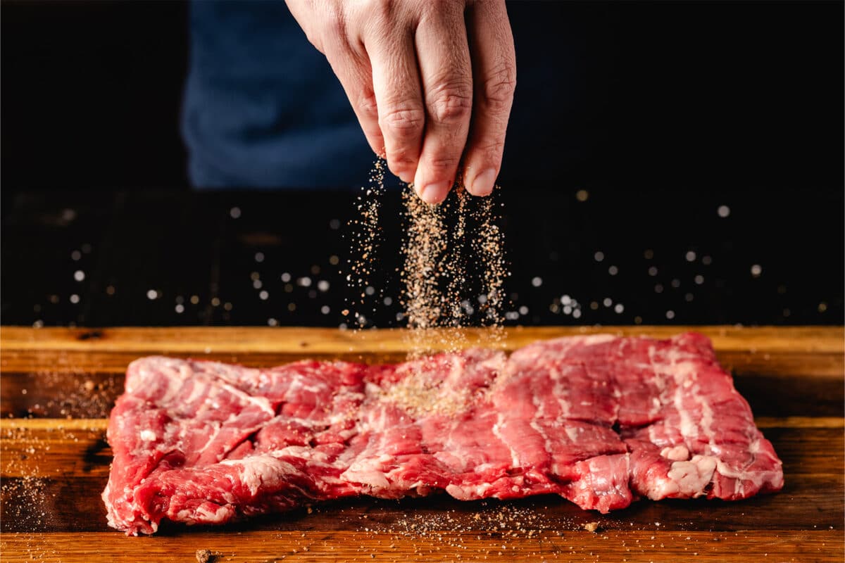 A raw skirt steak atop a wooden surface being seasoned with rub.